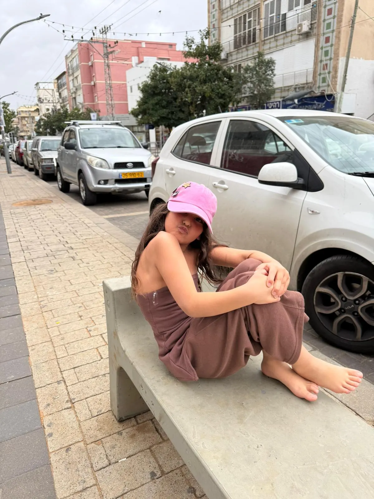 חליפת סטרפלס נויה שוקו בצבע חום שוקולד עם כובע ורוד, יושבת על ספסל בטון ליד רכב לבן, מבט מהצד